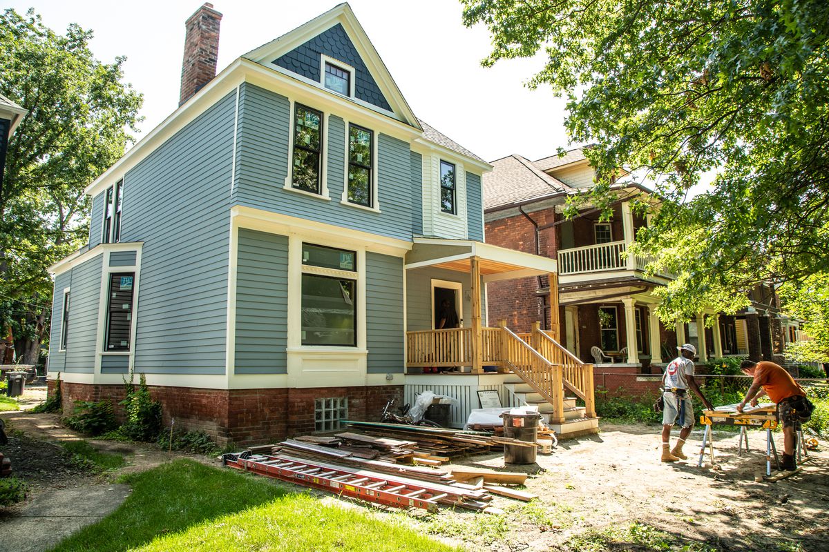 Two-story home with blue siding and a wood porch. Two carpenters saw wood in the front yard.
