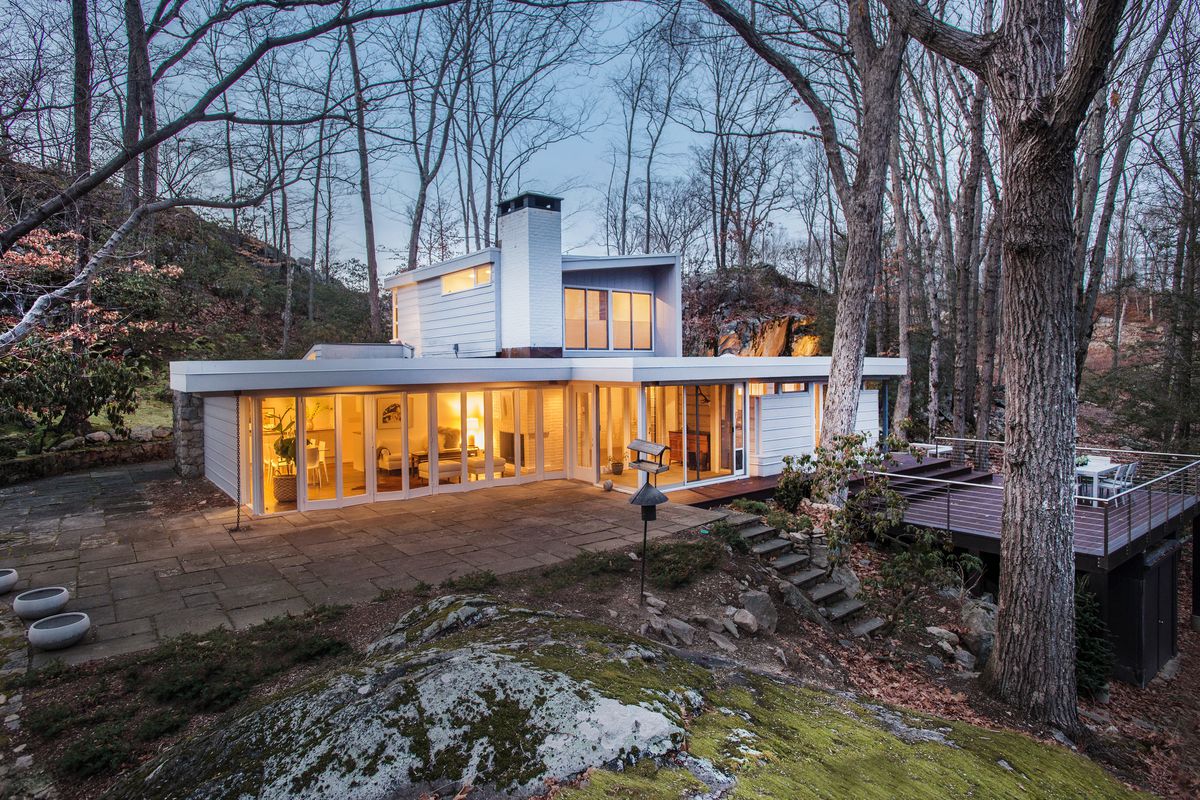 Exterior shot of white home with broad first floor and a smaller volume perched on top. It sits on a moss-covered hill in winter, with walls of accordion glass and a deck facing toward nature.  