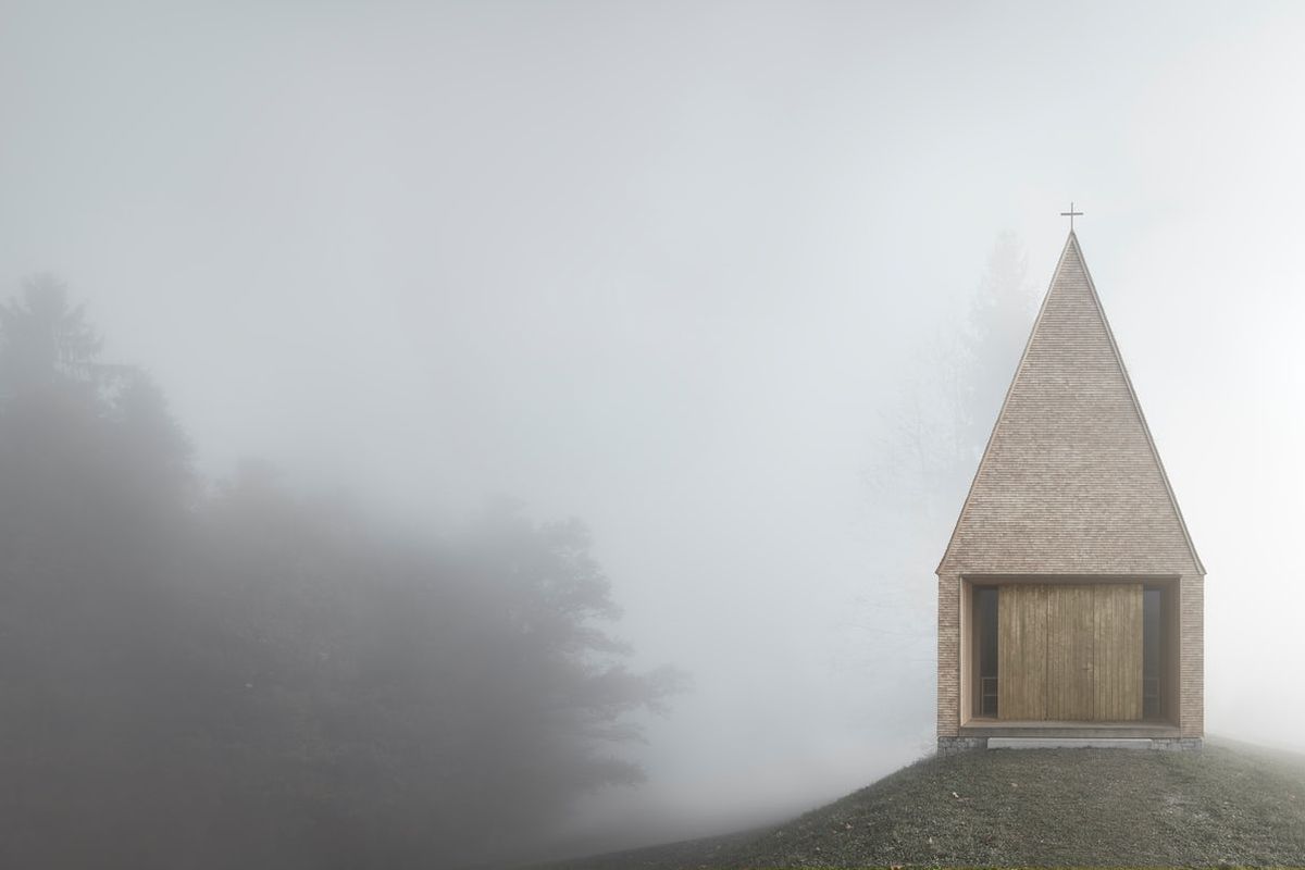 A small, simple chapel with high pitched roof all clad in wood sits atop a hill in haze.