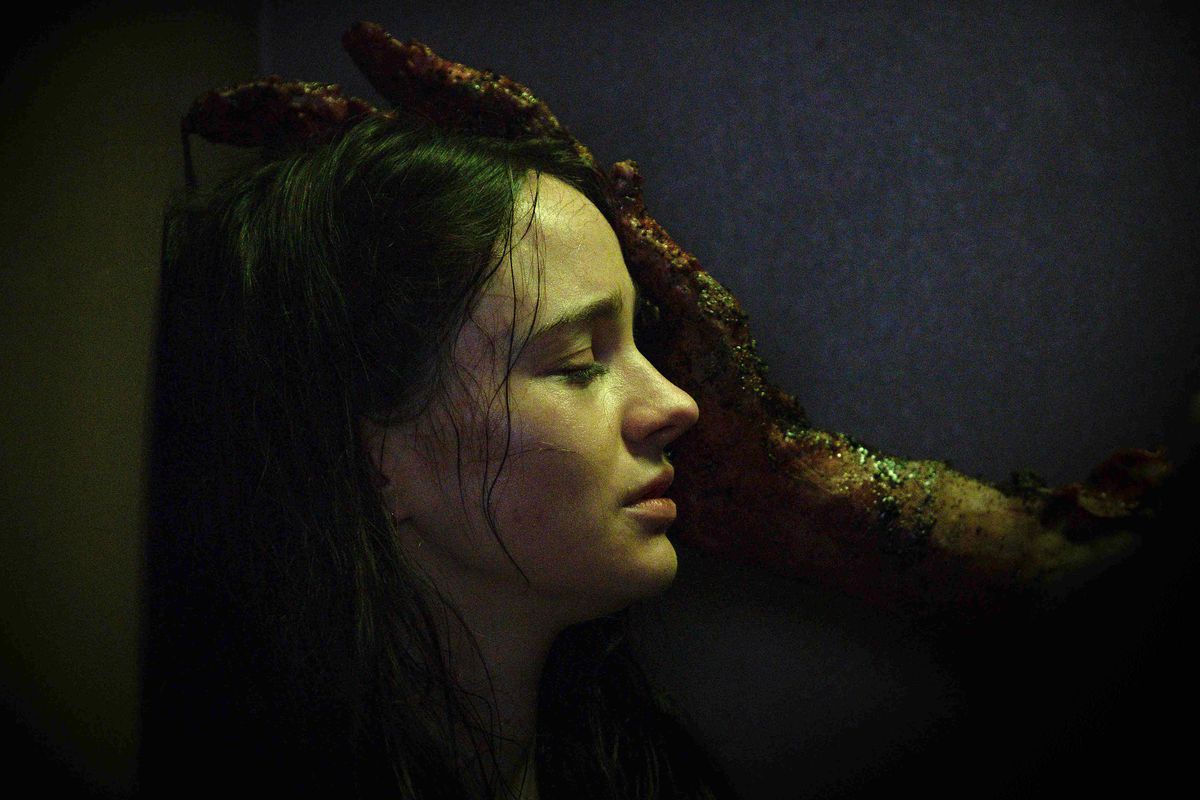 Ella (Aisling Franciosi), a young, pale woman with dark hair, closes her eyes and leans against a wall, looking like she’s close to crying, as a wet, gristly, dripping, inhuman hand reaches from offscreen to touch her face in the horror movie Stopmotion