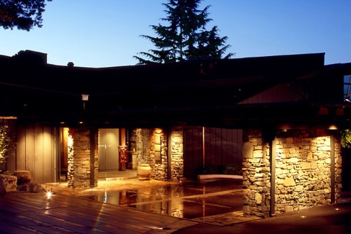 An exterior view of Canlis restaurant at night, with stone walls lighted in the foreground and the tops of trees seen in the background.