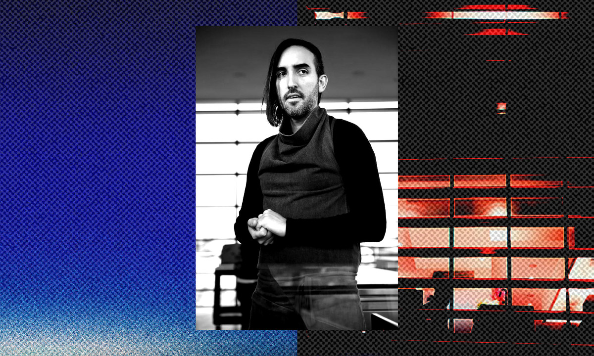 A black-and-white photograph of Jordan Kahn standing with his hands clasped together, against a graphic, pixelated background that is blue on one side and red and black on the other.