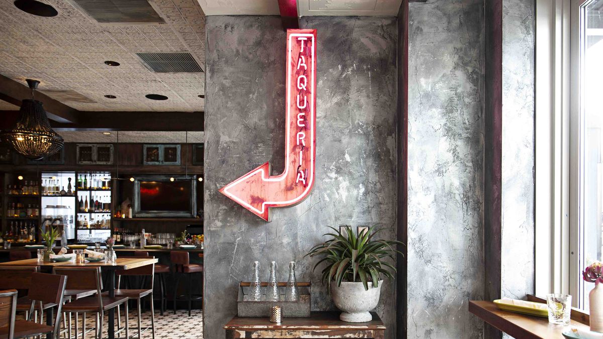 A red neon sign shaped like an arrow reads “taqueria” and hangs on a rustic gray wall, pointing toward a restaurant dining area