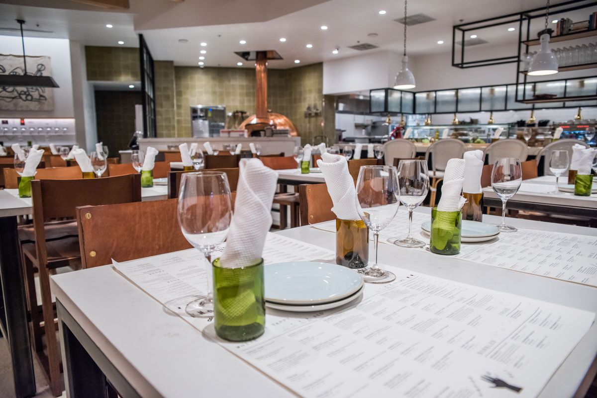 A tasting room and restaurant with white balls and mossy green tiles is photographed. In close relief is one table set with menus, wine glasses, water glasses made from wine bottles, napkins, and plates.