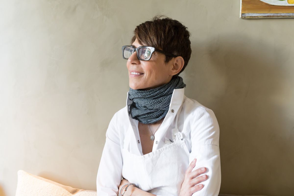 Chef Dominique Crenn wearing a scarf and glasses sitting in front of a cream-colored wall looking into the distance.