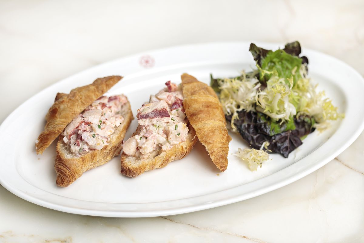 Two croissants sit on a white oval plate, each split open to reveal lobster salad. There’s also a small frisee salad on the plate. The plate sits on a white marble countertop.