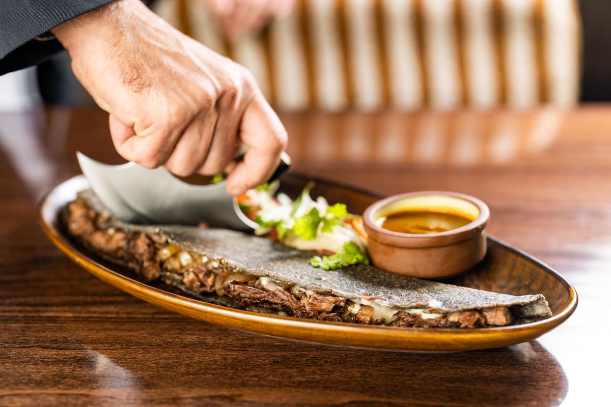 A machete, a football shaped Mexican street food like a quesadilla, sits on a brown plate with a bowl of dipping sauce. A man’s hand cuts it with a circular blade.