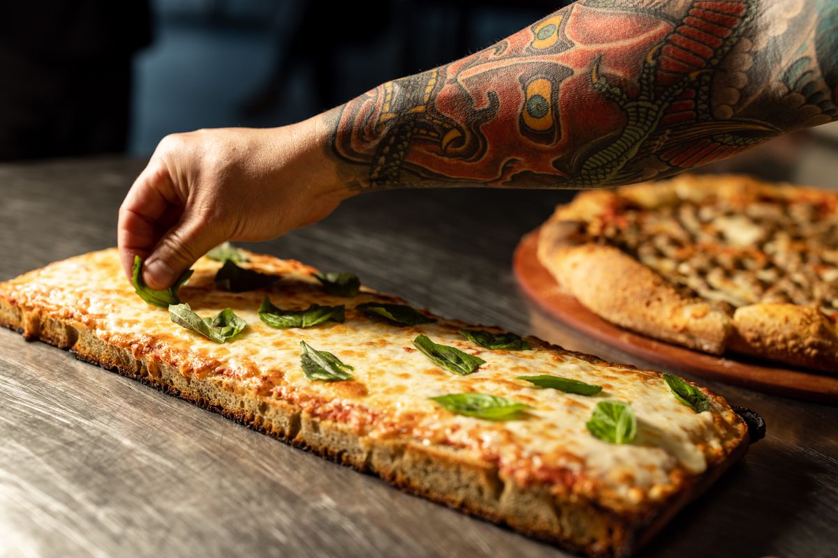 A man’s hand places basil on a rectangular pizza covered in cheese. 