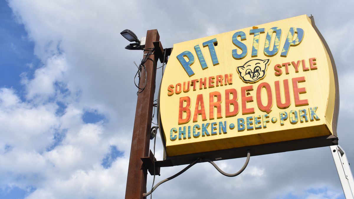 A faded restaurant sign is visible against a blue sky full of puffy white clouds. The sign says Pit Stop Southern Style Barbeque Chicken Beef Pork, and there’s a cartoon image of a pig’s face