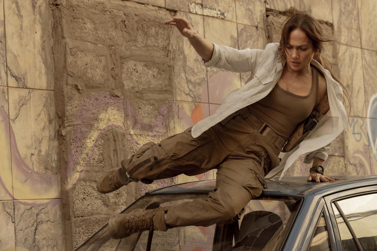An image of Jennifer Lopez in muted brown clothing and a white jacket sliding over the top of a car athletically.