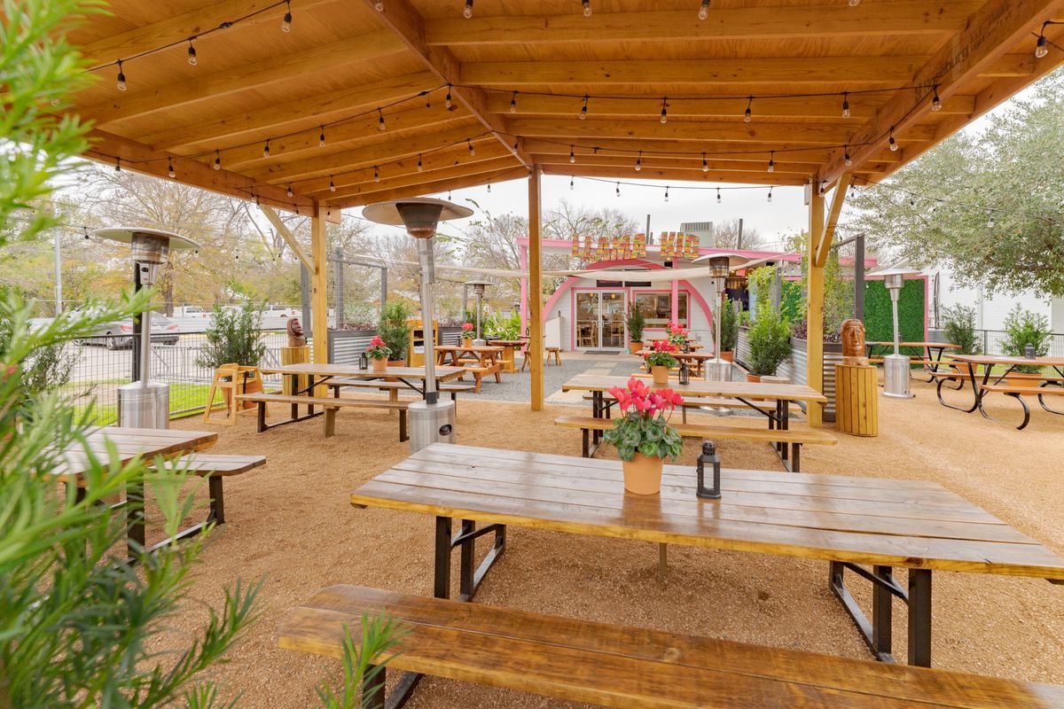 A restaurant patio with wooden picnic tables and benches with flower pots on each table underneath a wooden awning