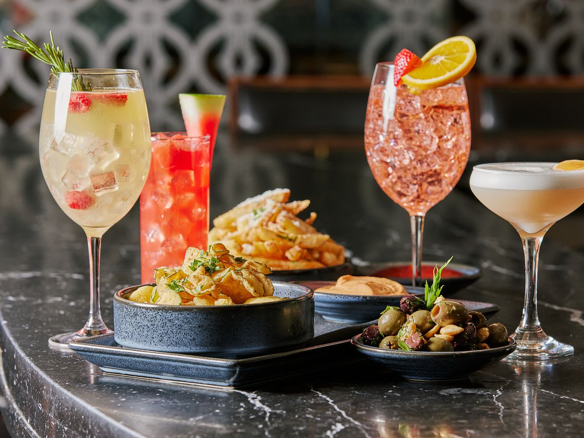 Plates of olives and fried snacks sit on a black marble bar top surrounded by cocktails.
