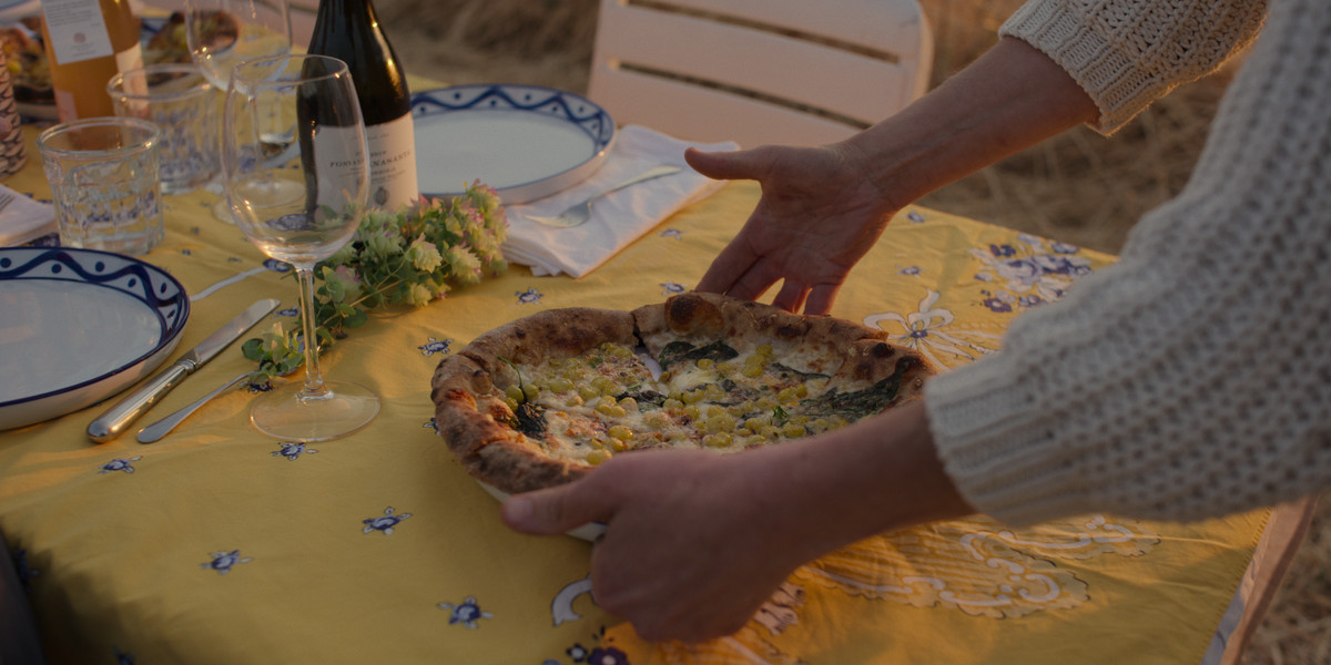 Two hands drop a corn-topped pie at a tablecloth-lined table in the garden.