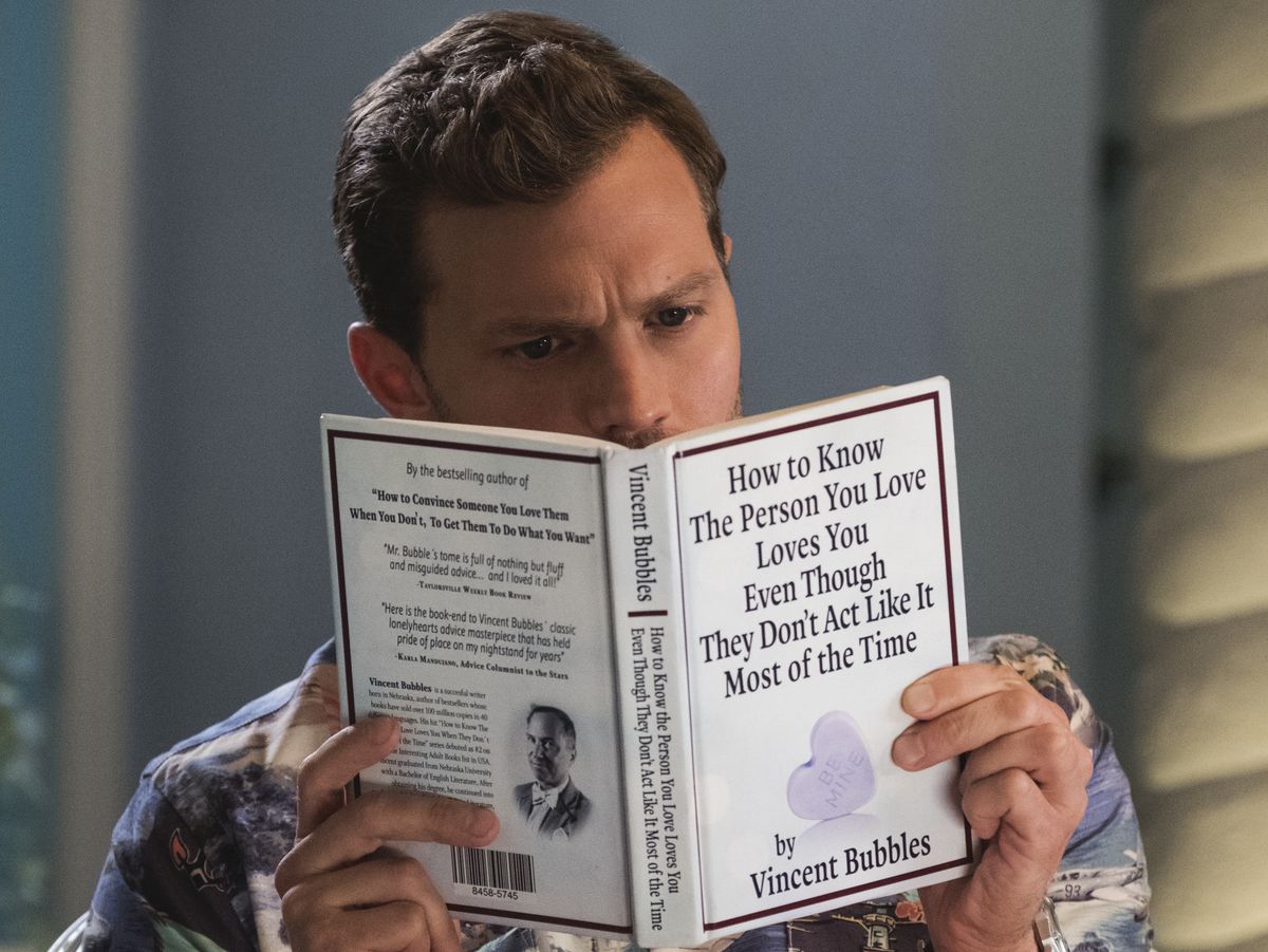 Jamie Dornan as Edgar reading a book called How to Know the Person You Love Loves You Even Though They Don’t Act Like It Most of the Time in Barb &amp; Star Go to Vista Del Mar