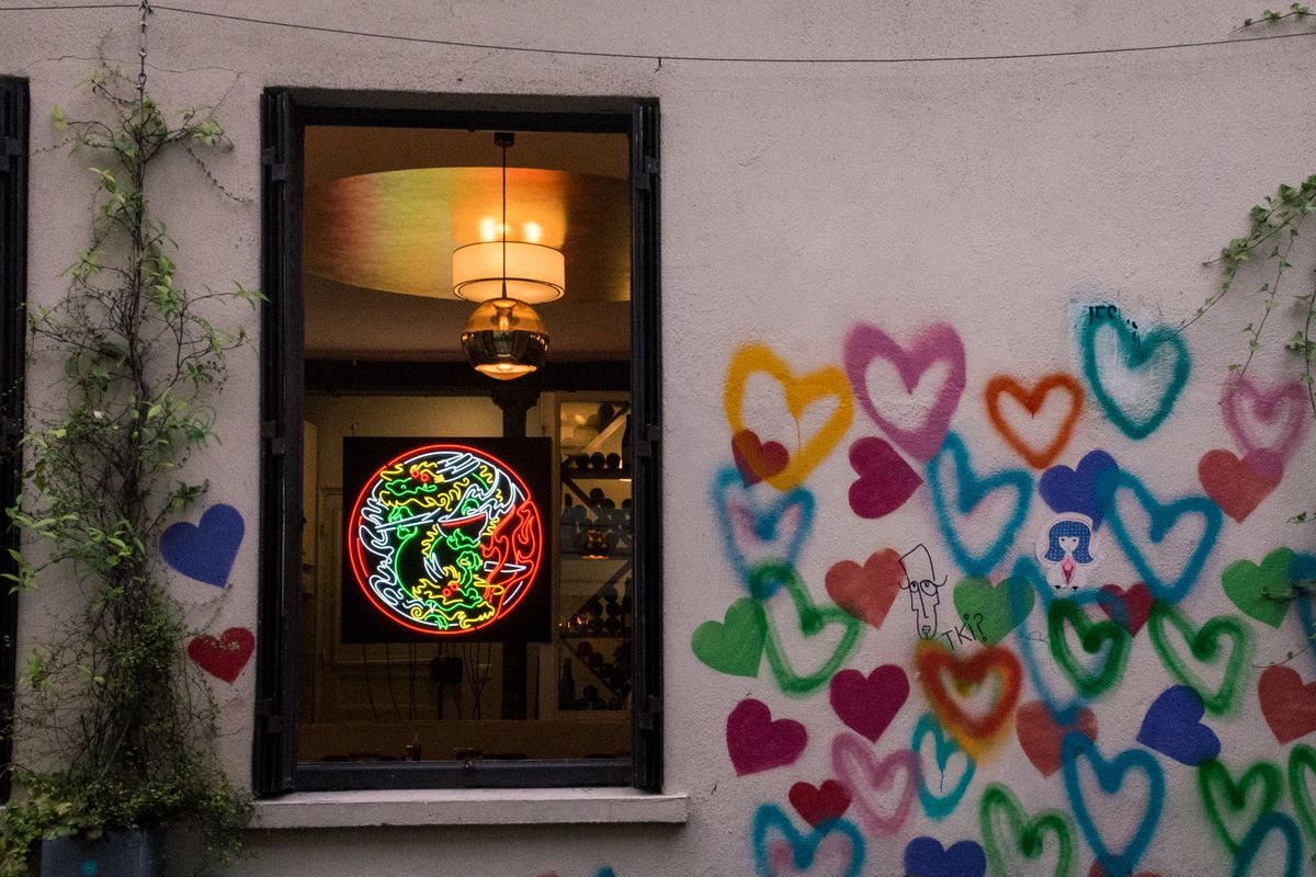 A white wall covered in graffiti of rainbow-colored hearts. A circular neon sign depicting an Asian dragon is visible through a window. 