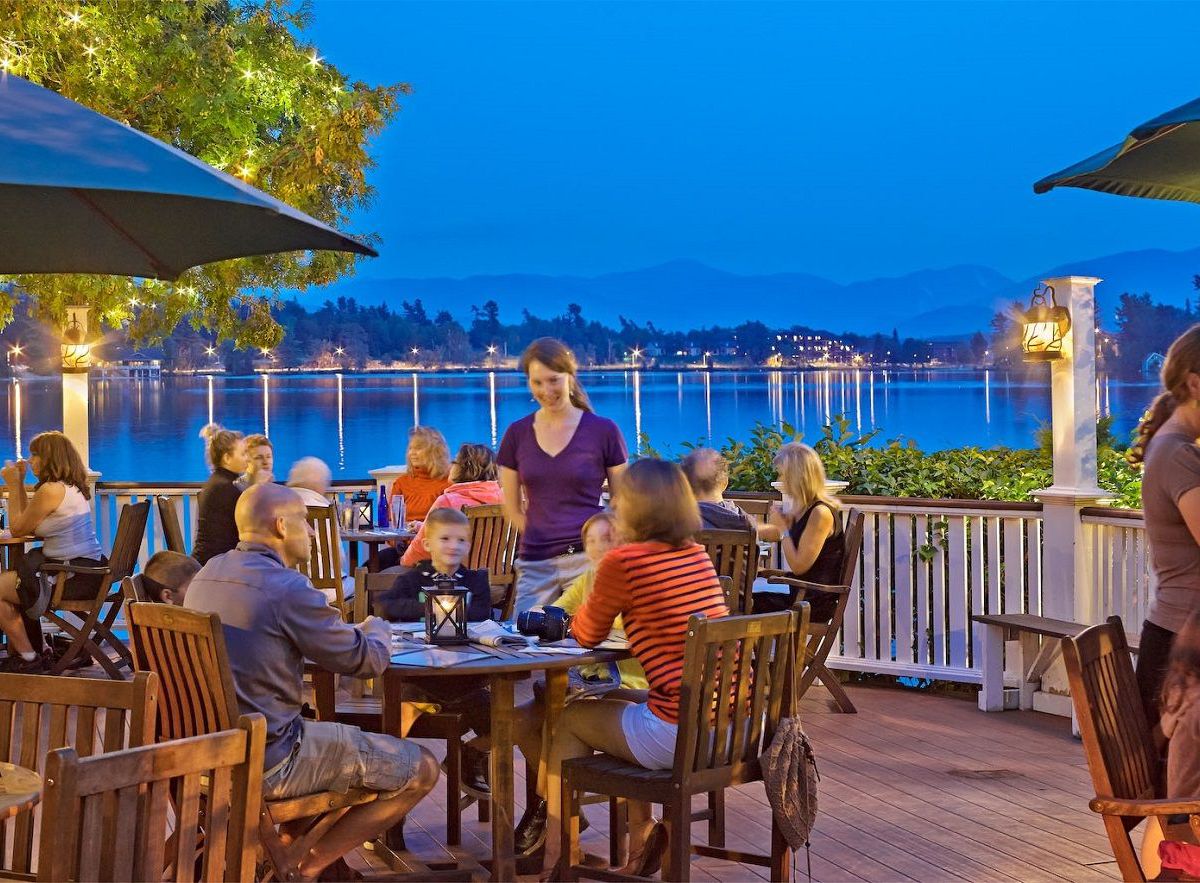 A waitress wearing a purple shirt stands besides a table of four diners, out on an outdoor dining patio with umbrellas. It is dusk, and behind the diners are views of Lake Placid and the panorama behind it. 