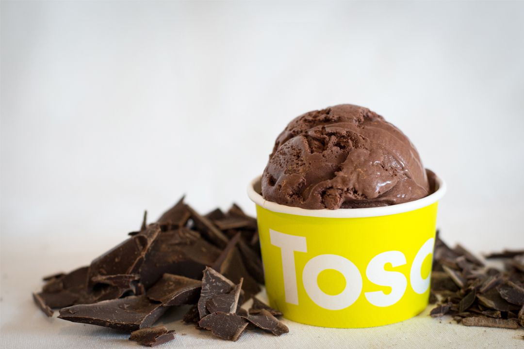 A scoop of chocolate ice cream in a branded yellow cup from Toscanini’s, with chunks of chocolate scattered in the background