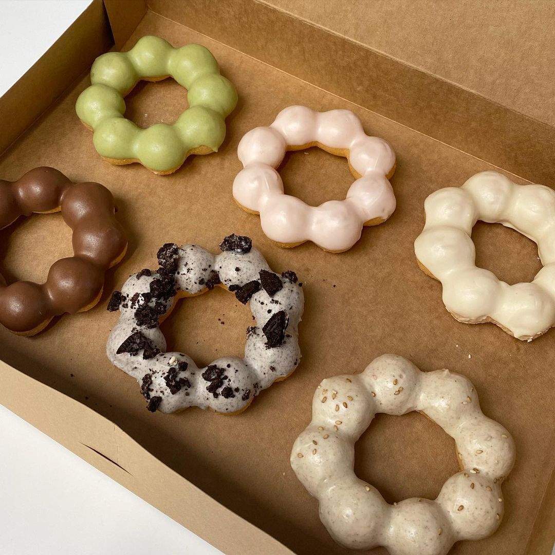 Six mochi doughnuts in six different flavors, each shaped like a ring of balls stuck together, are in a cardboard box.