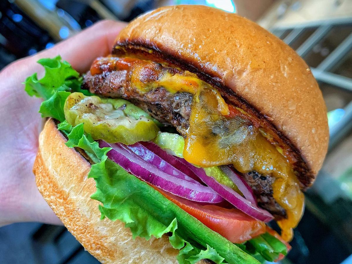 A hand holds a burger with cheese, pickles, onions, tomatoes, and lettuce on a bun