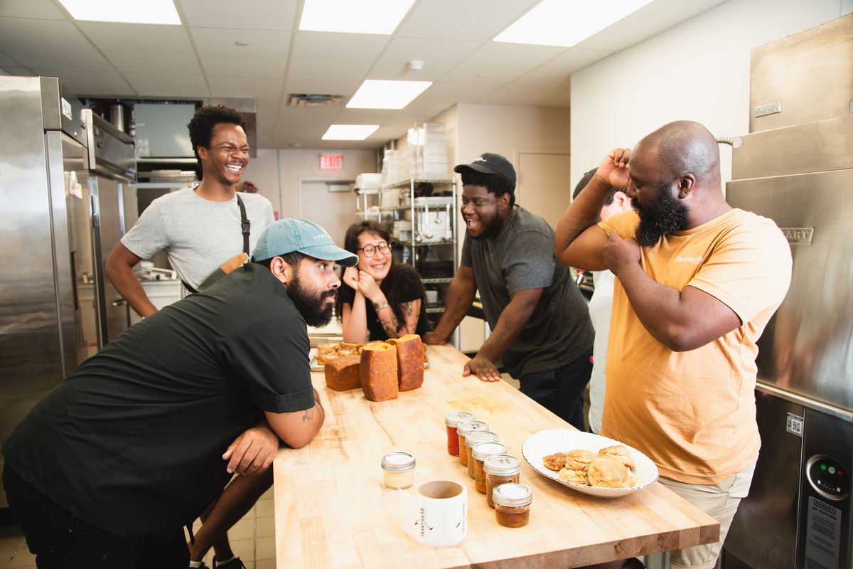 A group of chefs stand around a butcher block while one chef flexes his muscles and others laugh.