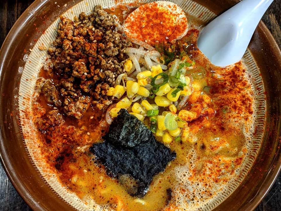 A brown bowl filled with miso ramen, sprinkled with red spice, corn, and a soft egg, with a white spoon resting in the bowl