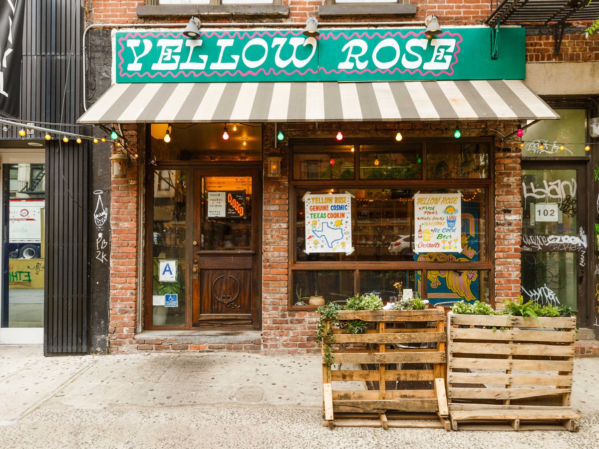 The sign for Yellow Rose, a mix of white lettering, pink bordering, and a teal background hangs above the striped awning of the venue