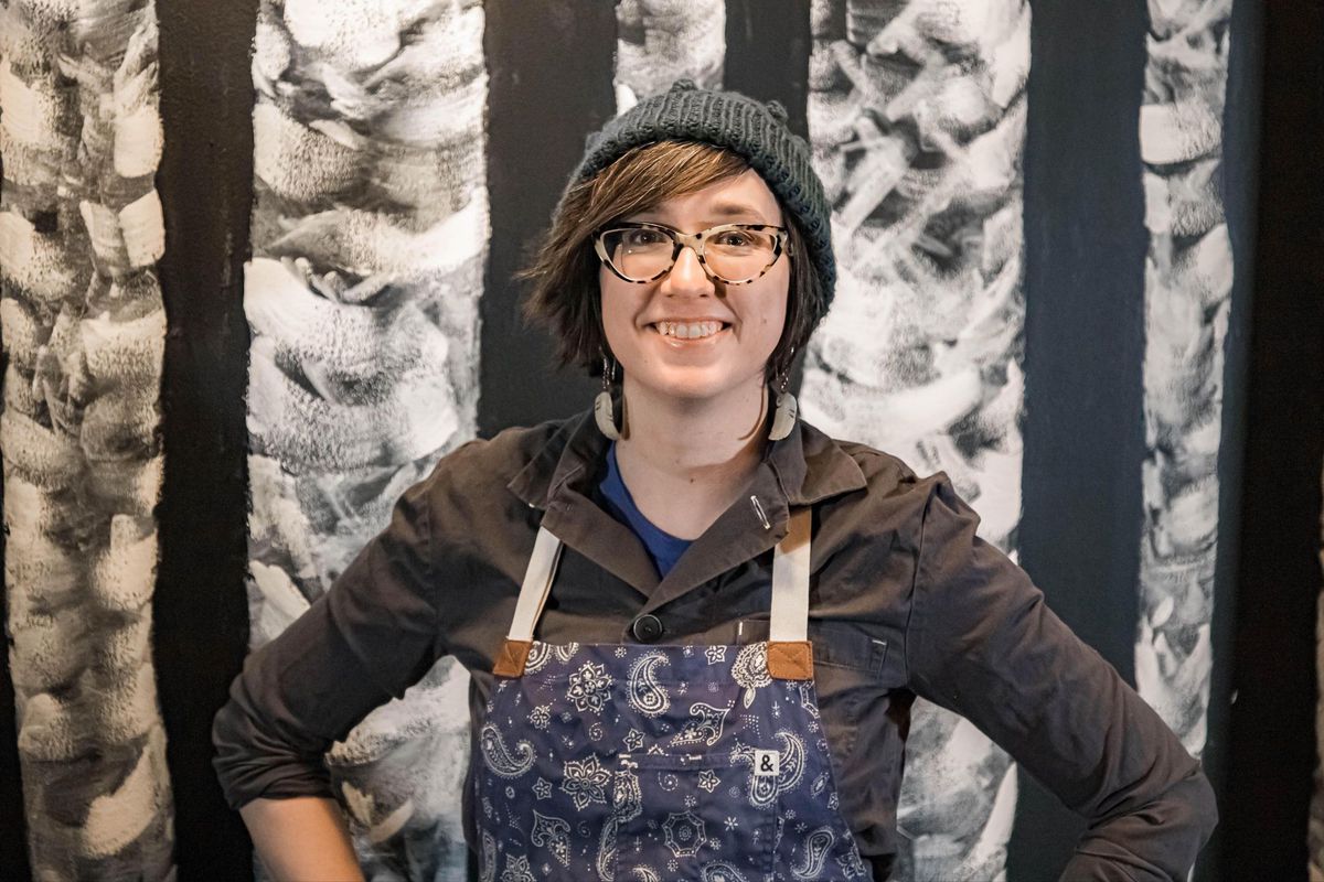 A woman with short brown hair, glasses, and a grey beanie wears an apron over a dark collared shirt and stands with hands on hips, smiling at the camera.