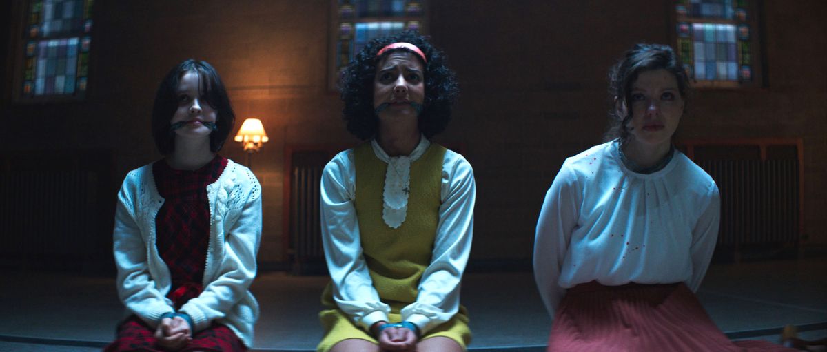 Clara (Georgia Acken) and Samantha (Madison Baines) kneel with their hands bound in front of them and gags in their mouths, next to their teacher Rose (Chloë Levine), kneeling with her hands bound behind her back in The Sacrifice Game