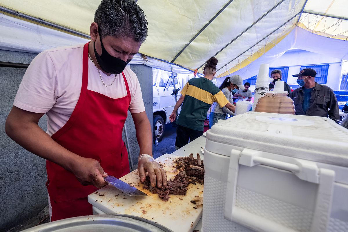 Julio James of Taxco Gro. cutting barbacoa de chivo at his stand in Los Angeles wearing a red apron.