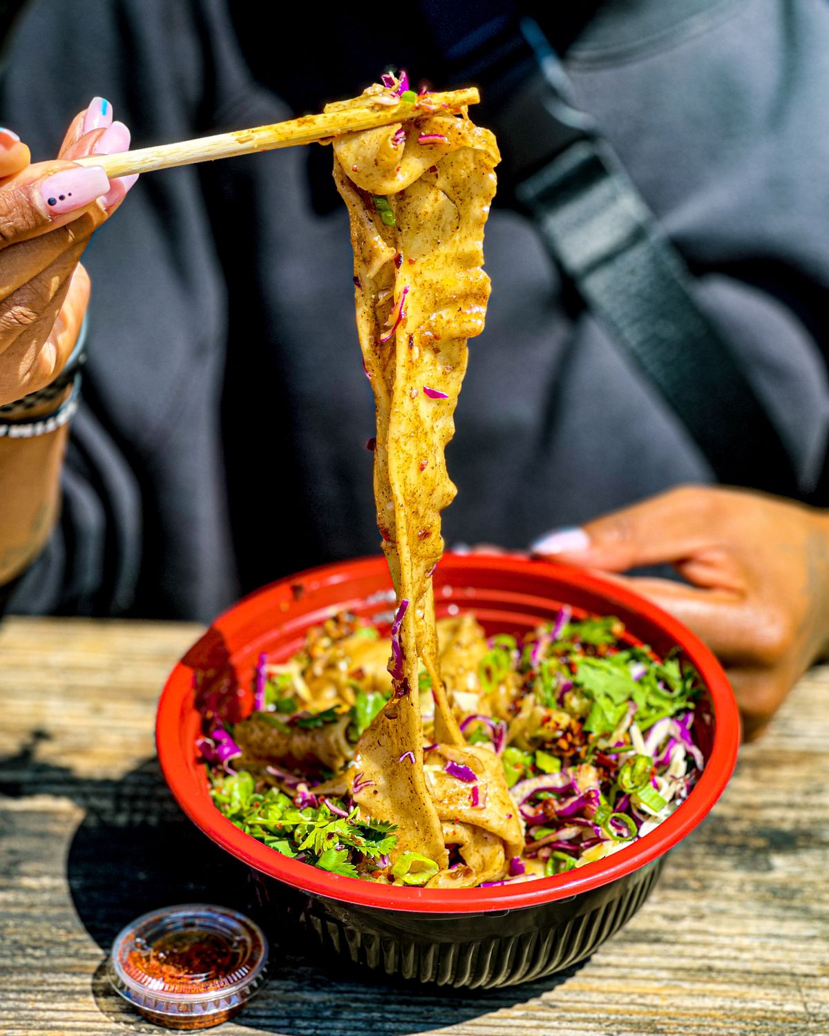 A hand with pink nails pulls thick noodles using chopsticks from a dark red bowl.