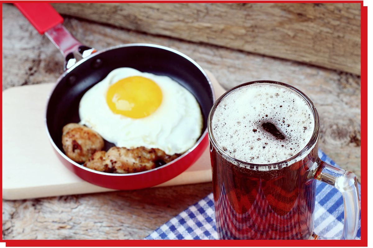 Pint of beer next to a small skillet containing fried egg and breakfast sausage.