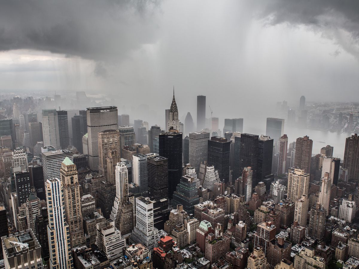 An aerial view of the buildings of New York City in midtown Manhattan. There are buildings of varying heights including the Chrysler Building. There are storm clouds and fog.