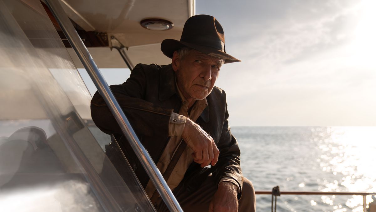Harrison Ford as Indiana Jones, wearing his trademark fedora and jacket, leaning on one knee on the side of a boat with the afternoon sun in the distance.