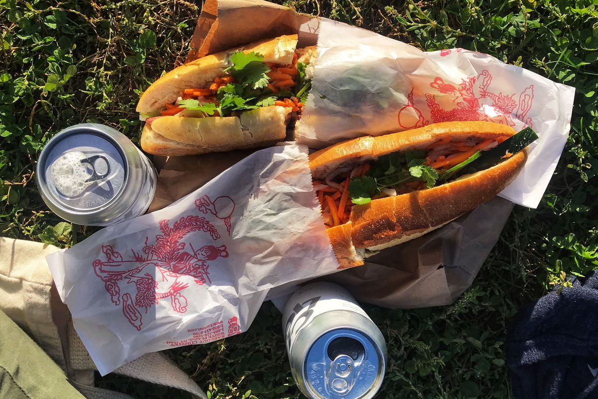 An overhead photograph of banh mi sandwiches and canned beers sitting in grass.