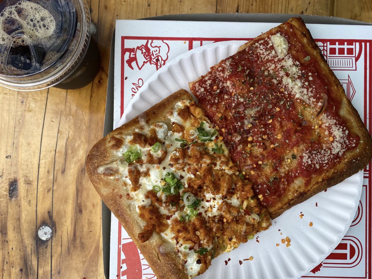 A square slice of buffalo chicken pizza, topped with mozzarella and green scallions, sits next to a tomato slice over a pizza box, next to a plastic cup of iced coffee