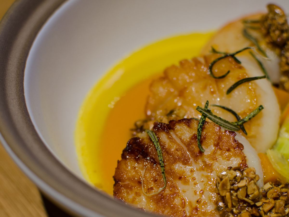 Seared scallops sit in a bowl on circles of orange and yellow purees, topped with herbs.