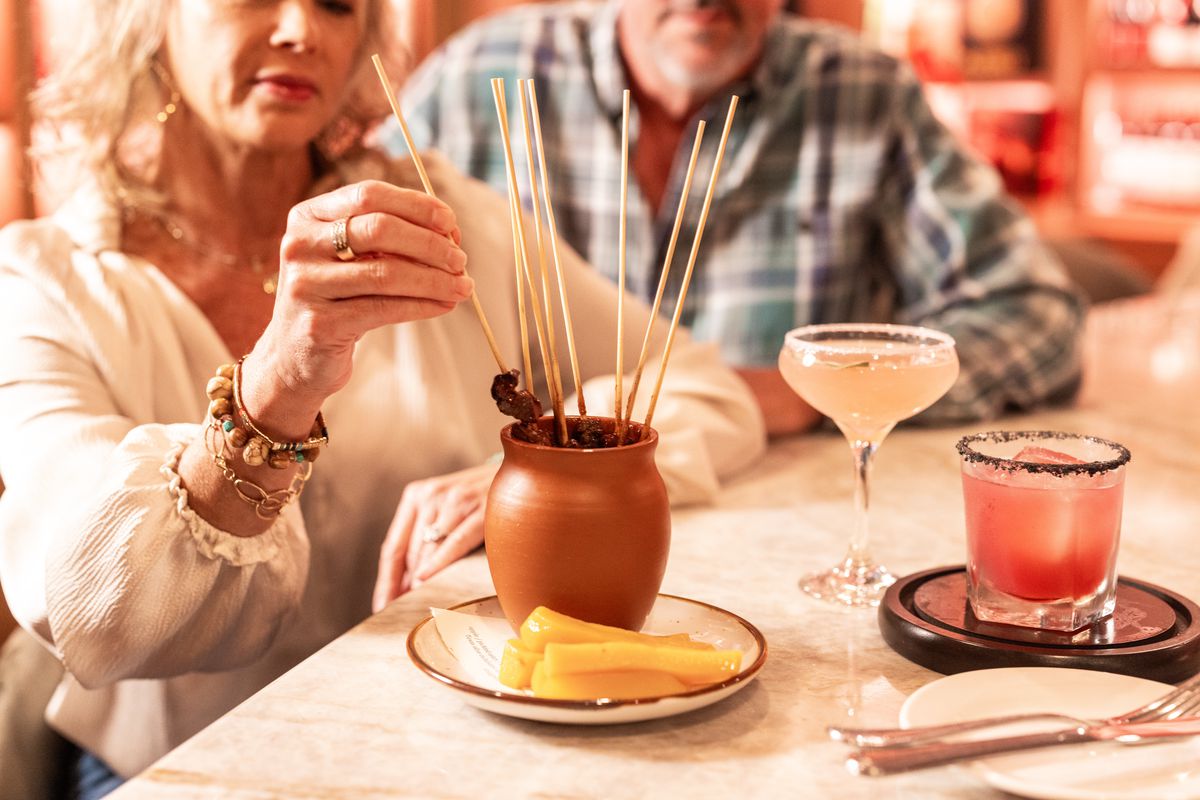A man and woman sit at a bar with cocktails in front of them. They’re removing chicken skewers from a clay pot to eat.