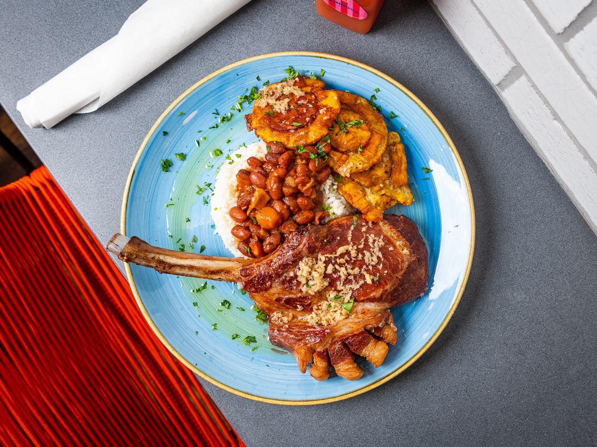 A blue plate holds a smashed, fried circles of plantain tostones, rice, pink beans, and a chuleta kan-kan (bone-in pork chop) that features a “mohawk” of pork belly cut to expose three edges from each fatty piece.