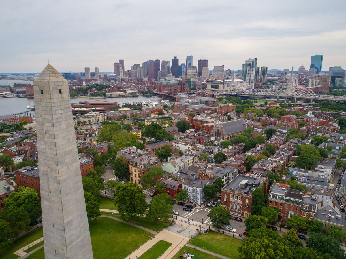An aerial view of city buildings in Boston. In the foreground is a tall monument. In the distance is a body of water and a skyline with many buildings.
