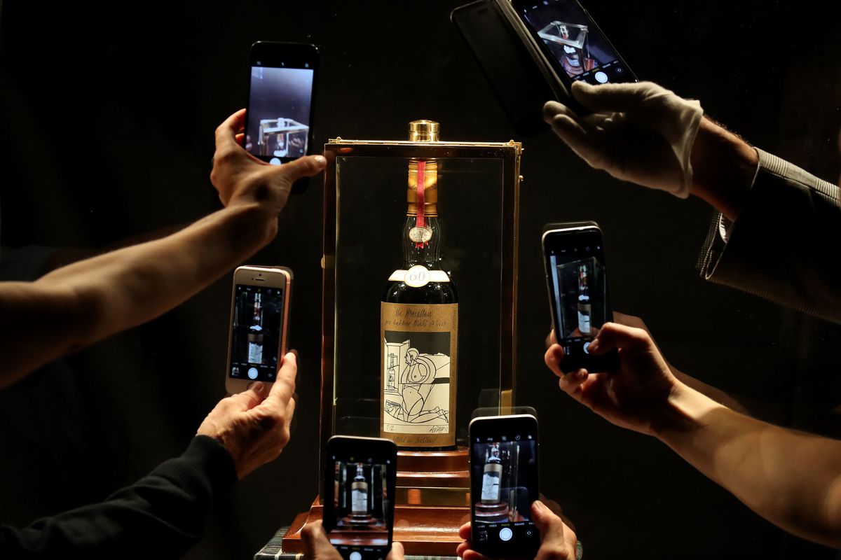 Hands holding cellphones up to take a picture of a bottle of whiskey in a display case.