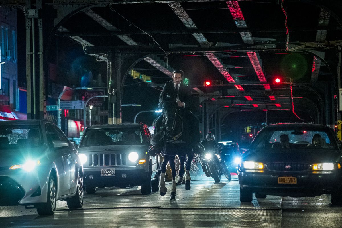 Keanu Reeves as John Wick, riding a horse under elevated train tracks at night, with a motorcycle chasing him through traffic