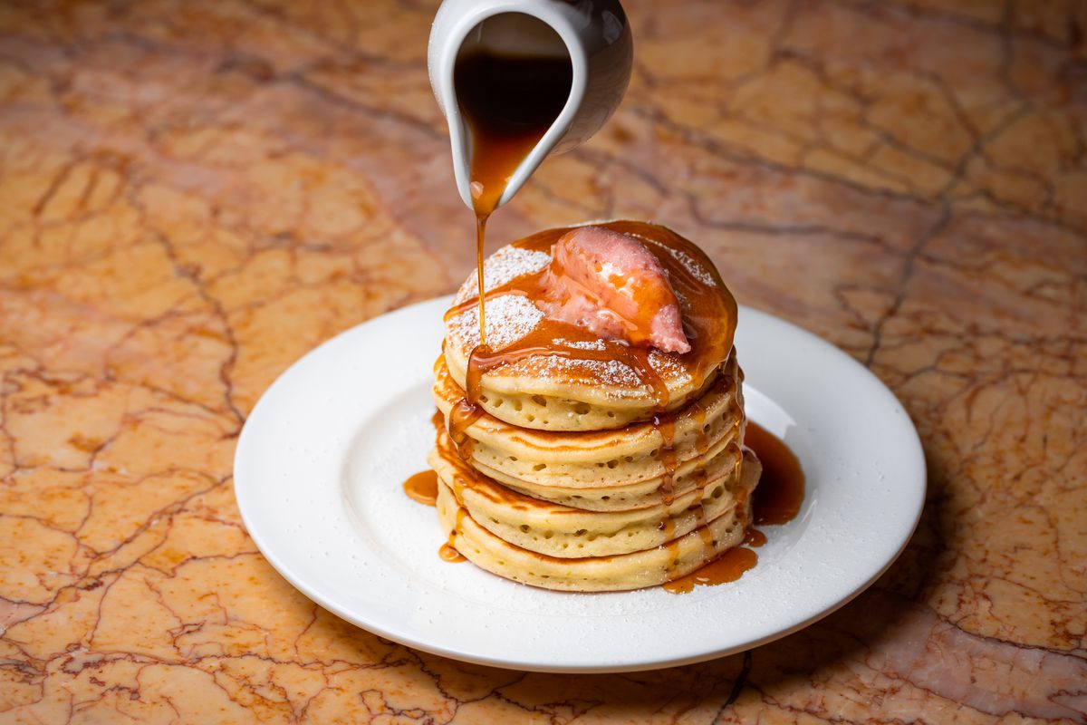 Syrup pours down on a narrow, tall stack of pancakes on a white plate on a brown table at modern restaurant Denae’s.