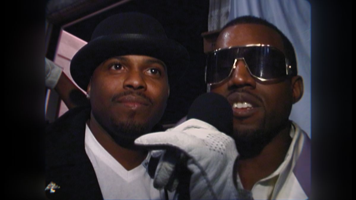 an image from the first episode of jeen-yuhs: A Kanye Trilogy of Kanye West, wearing sunglasses and holding a microphone with a white glove, standing next to another Black man wearing a fedora