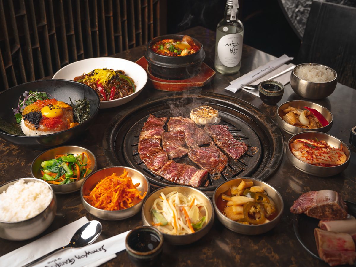 A table filled with dishes surrounding a tabletop grill with meat on it.