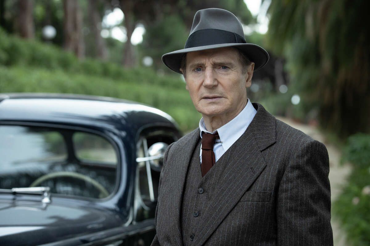 A man (Liam Neeson) standing in a forested area in front of a dark sedan dress in a brown pinstripe suit, dark red tie, and a gray fedora.