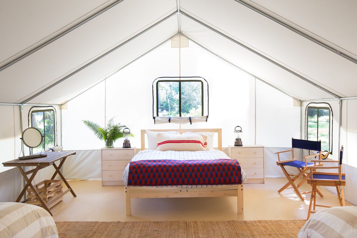 The interior of a large white tent. There is a bed on a bed frame, chairs, a table with a mirror, and plants in planters. The floor is wooden. There are windows in the walls of the tent. 