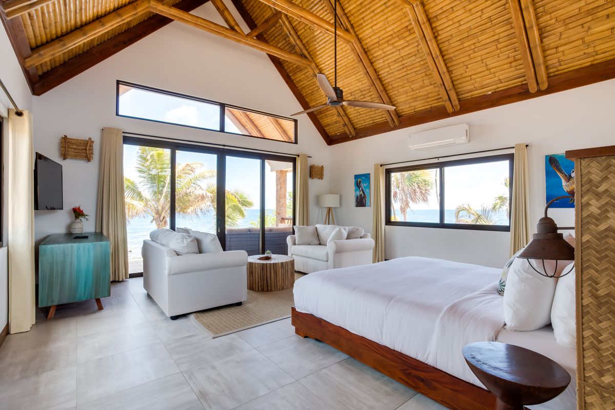 Interior of bedroom with pitched bamboo ceiling and windows with palm tree and ocean views. The room features a bed, two armchairs facing each other, and a TV mounted on the wall.