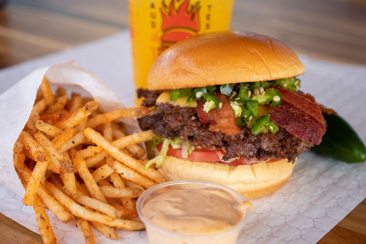 A burger with a meat patty, bacon slices, tomato slices, and diced jalapenos next to a white packet of spiced fries and in front of a yellow drink.
