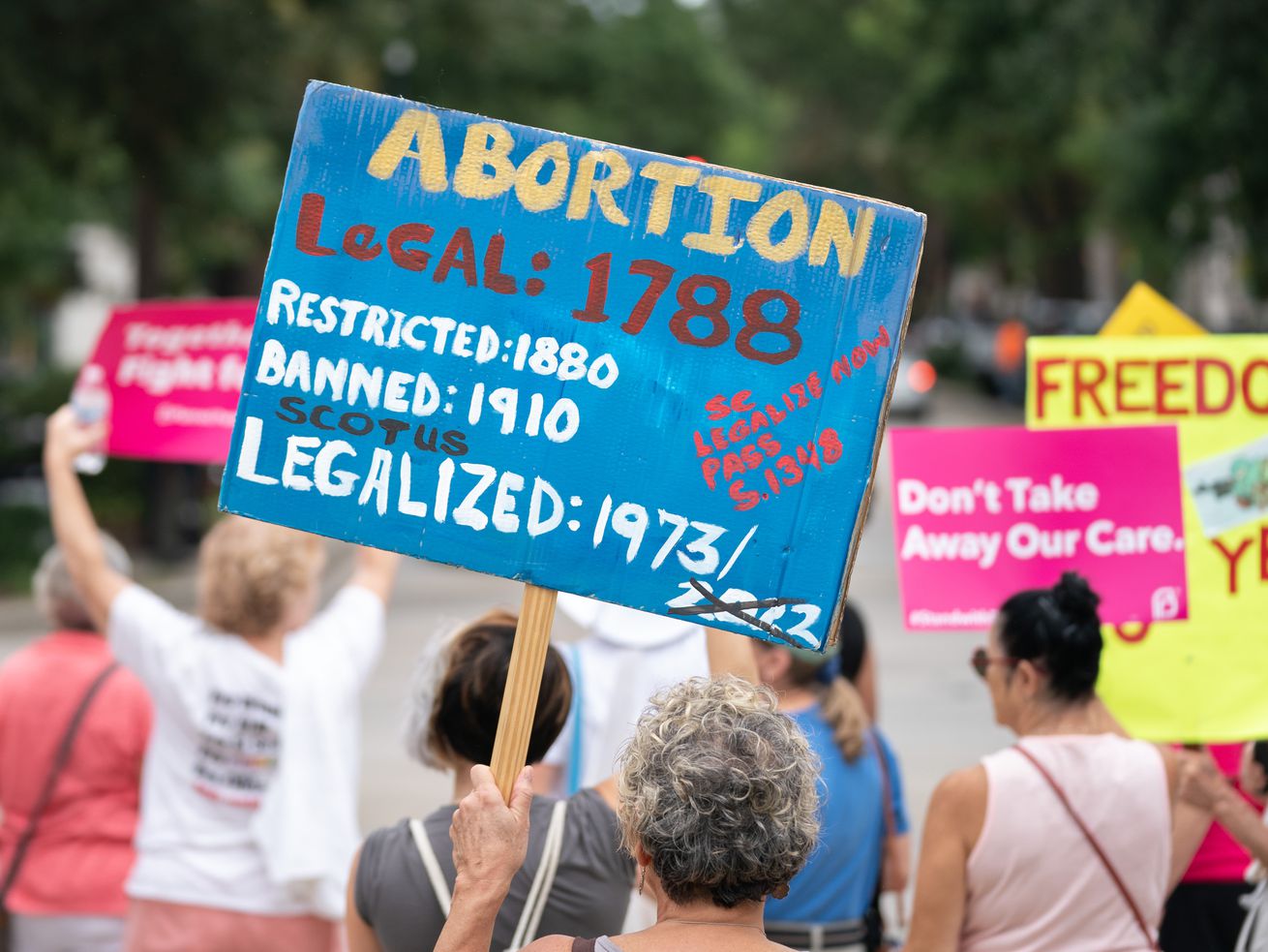 Protesters hold signs in front of the South Carolina Statehouse. Handpainted words on a large blue sign say: Abortion, legal: 1788, restricted: 1880, banned: 1910, legalized: 1973, with the year 2020 crossed out in black paint.