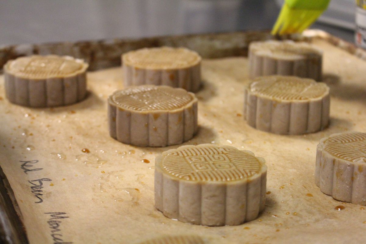 Round, unbaked mooncakes on a baking tray with parchment paper. Mooncakes are about an inch tall and 2 inches across in diameter, flat on top with a Taoist design stamped on top. A green pastry brush brushes molasses on top. The words “red bean mooncakes” is written at the bottom of the parchment paper.